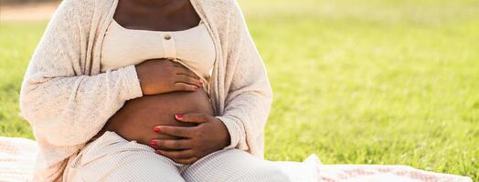 fechar acima grávida barriga do jovem africano mulher sentado dentro parque - maternidade estilo de vida conceito foto