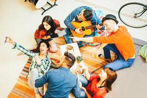 grupo do engraçado amigos desfrutando juntos jogando música com guitarra e levando selfie com Móvel telefone - feliz jovem pessoas tendo Diversão dentro a vivo quarto às casa - conceito do amizade foto