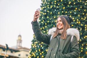 lindo jovem mulher levando uma selfie com Natal árvore atrás dela com Móvel telefone Câmera - bonita menina desfrutando Natal feriados levando uma auto fotografia dentro a cidade com dela Smartphone foto
