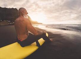 feliz em forma Senior homem sentado em prancha de surfe assistindo pôr do sol Tempo - maduro barbudo surfista tendo Diversão em surfar dia - extremo esporte e saúde pessoas estilo de vida conceito foto