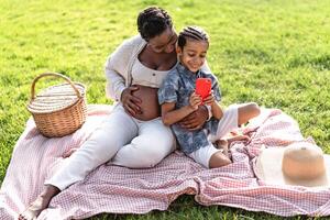 feliz africano grávida mãe gastos Tempo com dele filho fazendo uma piquenique durante final de semana dentro público parque - afro família assistindo em Smartphone - maternidade e pais estilo de vida conceito foto