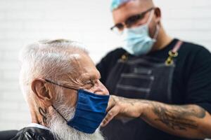 masculino cabeleireiro corte cabelo para hipster Senior cliente enquanto vestindo face cirúrgico mascarar - jovem estilista de cabelo trabalhando dentro barbearia durante corona vírus surto - saúde Cuidado e corte de cabelo salão conceito foto