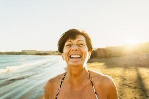 feliz caucasiano Senior mulher tendo Diversão sorridente para dentro a Câmera em a de praia durante verão período de férias - idosos pessoas estilo de vida conceito foto