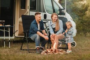 uma família cozinheiros salsichas em uma fogueira perto seus motorhome dentro a madeiras foto