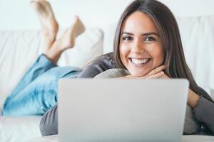 jovem mulher deitado em sofá e usando computador portátil às casa - feliz menina surfar conectados com dela computador enquanto sorridente às Câmera - conceito do pessoas, tecnologia e social meios de comunicação redes foto