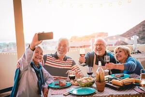 feliz idosos amigos levando selfie com Móvel Smartphone Câmera às churrasco jantar - maduro pessoas tendo Diversão comendo e bebendo vermelho vinho em pátio enquanto usando Novo tendências tecnologia telefone apps foto