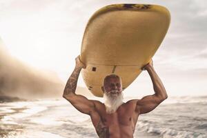 feliz em forma Senior tendo Diversão surfar às pôr do sol Tempo - desportivo barbudo homem Treinamento com prancha de surfe em a de praia - idosos saudável pessoas estilo de vida e extremo esporte concep foto