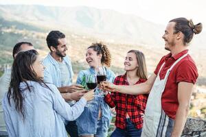 grupo do feliz amigos torcendo e brindar com vermelho vinho óculos em terraço - jovem pessoas tendo Diversão bebendo às jantar festa em pátio com panorâmico Visão - amizade e juventude estilo de vida conceito foto