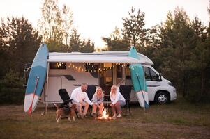 uma família cozinheiros salsichas em uma fogueira perto seus motorhome dentro a madeiras foto