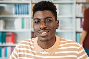 feliz jovem africano homem sorridente para dentro a Câmera enquanto em pé dentro universidade biblioteca foto