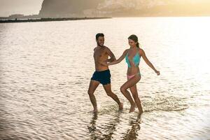 jovem feliz casal tendo Diversão corrida dentro a mar segurando seus mãos às pôr do sol - romântico e concurso momentos do amantes dentro período de férias em a de praia - amor, relação, viagem conceito foto