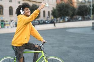 feliz africano homem levando selfie com Móvel Smartphone ao ar livre - jovem cara tendo Diversão equitação com bicicleta dentro a cidade - juventude milenar geração estilo de vida e tecnologia conceito foto