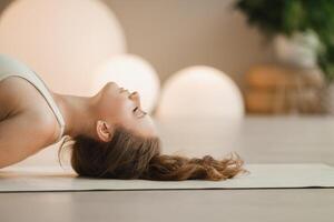 uma menina dentro branco roupas faz ioga deitado em uma tapete dentro de casa foto