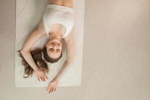 uma menina dentro branco roupas mentiras em uma tapete e risos antes ioga Aulas dentro de casa foto
