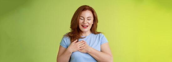 fascinado fofa ruiva apaixonado meia idade mulher suspirando adorável toque coração fechar olhos sorridente Deleitado expressar admiração tentação sentindo-me apreciação grato emoções verde fundo foto