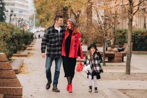 uma pequeno menina com dela pais corre para a outono cidade.a à moda família do três passeios através a outono cidade foto