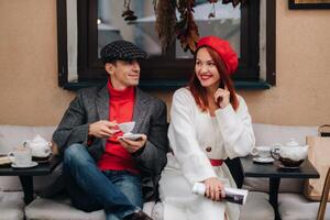 uma feliz à moda casal bebidas café e sorrisos enquanto sentado dentro uma cafeteria em a rua foto