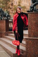 uma lindo à moda mulher vestido dentro a elegante vermelho casaco com uma à moda vermelho Bolsa dentro a outono cidade foto