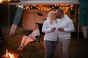 uma casado casal com óculos do vinho carrinhos contra a fundo do uma motorhome e descansos juntos de a fogueira. tarde família período de férias foto