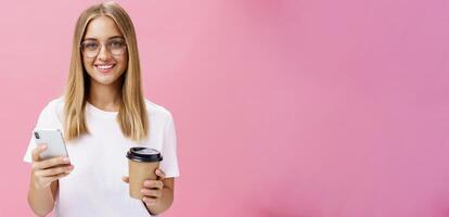 agradável de aparência amigável menina sorridente às Câmera segurando papel copo do café e Smartphone. retrato do alegre agradável mulher bebendo manhã bebida, posando opinião sobre cafeteria dentro Internet sobre Rosa parede foto