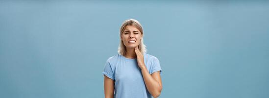 ai meu garganta. descontente infeliz boa aparência adulto Loiras menina com curto corte de cabelo aperto dentes a partir de doloroso sentindo-me tocante pescoço tendo angina ou inflamação posando sobre azul fundo foto