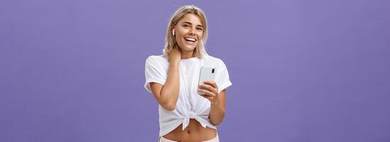 tímido atraente e à moda sociável mulher dentro branco camiseta tocante pescoço tímido e desajeitado sorridente amplamente vestindo sem fio fones de ouvido enquanto ouvindo canções com Smartphone sobre roxa parede foto