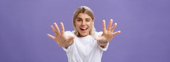 alcançando para você. retrato do provocante boa aparência e confiante à moda mulher com tatuagens em braços piscando degola Fora língua de brincadeira e sorridente puxar mãos para Câmera sobre roxa fundo foto