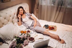 uma menina senta dentro cama dentro a tarde assistindo uma computador portátil e comendo morangos, uma menina dentro cama come doces antes indo para cama foto