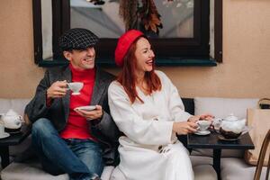 uma feliz à moda casal bebidas café e sorrisos enquanto sentado dentro uma cafeteria em a rua foto