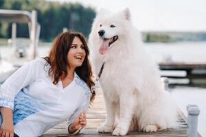 uma feliz mulher com uma grande branco cachorro mentiras em uma cais perto a mar às pôr do sol foto