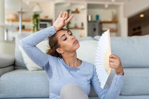 estressado irritado mulher usando acenando ventilador Sofra a partir de superaquecimento, verão calor saúde hormônio problema, não ar condicionador às casa sentar em sofá sentir exaustão desidratação insolação conceito foto