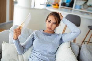 estressado irritado mulher usando acenando ventilador Sofra a partir de superaquecimento, verão calor saúde hormônio problema, não ar condicionador às casa sentar em sofá sentir exaustão desidratação insolação conceito foto