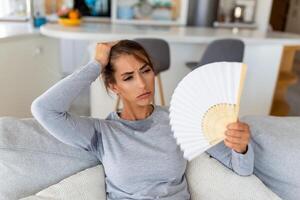 estressado irritado mulher usando acenando ventilador Sofra a partir de superaquecimento, verão calor saúde hormônio problema, não ar condicionador às casa sentar em sofá sentir exaustão desidratação insolação conceito foto