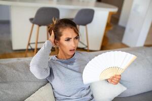 estressado irritado mulher usando acenando ventilador Sofra a partir de superaquecimento, verão calor saúde hormônio problema, mulher suor sentir desconfortável quente dentro verão clima problema sem ar condicionador foto