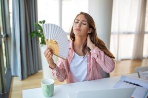 uma jovem mulher suores profusamente enquanto trabalhando em dela computador portátil dentro a superaquecido casa escritório. com não ar condicionador, ela se apega para uma onda fã, lutando a intenso verão aquecer. foto