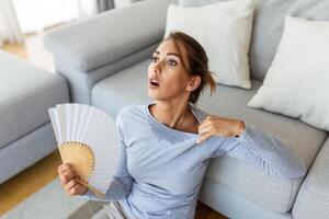 estressado irritado mulher usando acenando ventilador Sofra a partir de superaquecimento, verão calor saúde hormônio problema, mulher suor sentir desconfortável quente dentro verão clima problema sem ar condicionador foto