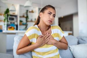 estressado jovem mulher sentindo-me dor e tocante peito Sofra a partir de mágoa doença às casa enquanto tendo coração ataque, Infarte foto