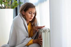 mulher congelando às lar, sentado de a frio radiador. mulher com casa aquecimento problema sentindo-me frio. poder restrição. foto