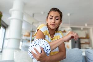 jovem mulher é sentado às casa em dela sofá e tocante a dela cotovelo enquanto sofrimento a partir de cotovelo dor, colocação gelo pacote em doeu local foto