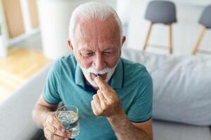 Senior homem leva comprimido com vidro do água dentro mão. estressado maduro homem bebendo sedado antidepressivo remédios. homem sente depressivo, levando drogas. medicação às trabalhos foto