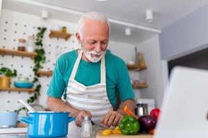 feliz Senior homem tendo Diversão cozinhando às casa - idosos pessoa preparando saúde almoço dentro moderno cozinha - aposentado estilo de vida Tempo e Comida nutrição conceito foto