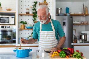 feliz Senior homem tendo Diversão cozinhando às casa - idosos pessoa preparando saúde almoço dentro moderno cozinha - aposentado estilo de vida Tempo e Comida nutrição conceito foto