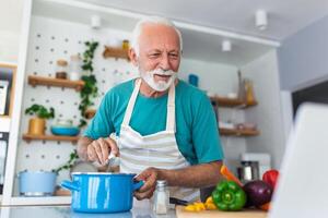 feliz Senior homem tendo Diversão cozinhando às casa - idosos pessoa preparando saudável almoço dentro moderno cozinha olhando às a recibo às dele computador portátil foto