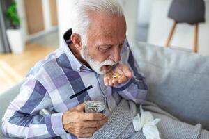 Senior homem levando pílulas enquanto sentado em sofá às lar, segurando branco jarra com tratamento, cópia de espaço. grisalho idosos homem usando suplementos ou vitaminas. saudável estilo de vida dentro Senior era conceito foto