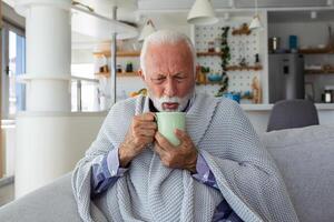 Senior homem sofrimento a partir de gripe bebendo chá enquanto sentado embrulhado dentro uma cobertor em a sofá às lar. doente Mais velho homem com dor de cabeça sentado debaixo a cobertor dentro a vivo sala. foto