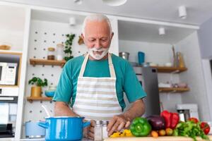 feliz Senior homem tendo Diversão cozinhando às casa - idosos pessoa preparando saúde almoço dentro moderno cozinha - aposentado estilo de vida Tempo e Comida nutrição conceito foto