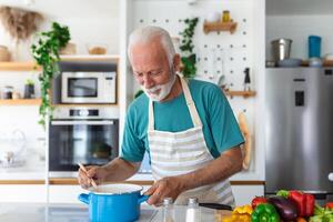 feliz Senior homem tendo Diversão cozinhando às casa - idosos pessoa preparando saúde almoço dentro moderno cozinha - aposentado estilo de vida Tempo e Comida nutrição conceito foto