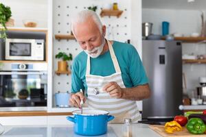 feliz Senior homem tendo Diversão cozinhando às casa - idosos pessoa preparando saúde almoço dentro moderno cozinha - aposentado estilo de vida Tempo e Comida nutrição conceito foto