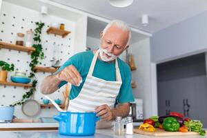 feliz Senior homem tendo Diversão cozinhando às casa - idosos pessoa preparando saúde almoço dentro moderno cozinha - aposentado estilo de vida Tempo e Comida nutrição conceito foto