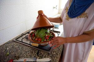 fechar-se meio Oriental dona de casa com cabeça coberto dentro hijab, em pé de forno, cozinhando marroquino tajine com legumes foto